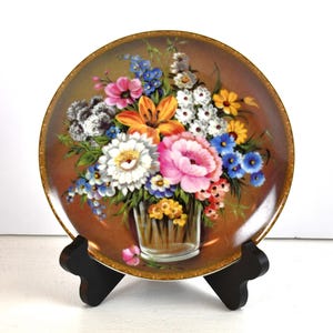 Peut inclure: Une assiette décorative avec une image peinte d'un bouquet de fleurs colorées dans un vase en verre transparent. Les fleurs comprennent des fleurs roses, blanches, jaunes, orange, bleues et violettes. L'assiette a un fond brun avec un bord doré.