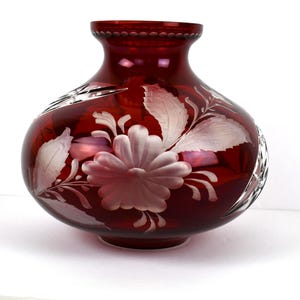 Puede incluir: Un jarrón de cristal rojo rubí con cuerpo bulboso y cuello acampanado. El jarrón presenta diseños florales grabados, incluyendo una flor grande y hojas. El cristal tiene un acabado brillante y está sobre una superficie blanca.