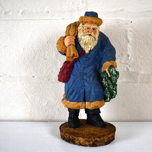 May include: A blue and brown wooden figurine of Santa Claus holding a bag of gifts and a sprig of holly. He is wearing a blue coat with a fur trim, a brown hat, and a white beard.