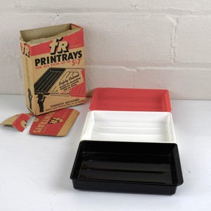 May include: A set of three rectangular print trays in red, white, and black, with a cardboard box labeled "Printrays" in red and black. The box is partially open, and the trays are stacked.