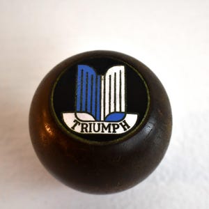 May include: A brown wooden gear shift knob with a black and white Triumph logo with blue accents.