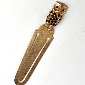 Könnte beinhalten: Ein goldfarbener Metall-Lesezeichen mit einem Eulen-Design. Die Eule hat schwarze, edelsteinartige Akzente auf Körper und Augen. Das Lesezeichen ist rechteckig mit einem dekorativen Oberteil und einem geschlitzten Design.