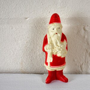May include: A vintage plastic Santa Claus figurine with a red suit, white beard, and a white hat. He is holding a bag of gifts in his left hand.