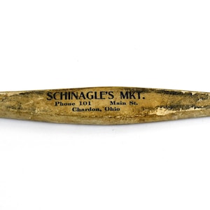 May include: A weathered, elongated wooden object with pointed ends. The text "SCHINAGLE'S MKT." is printed in black, along with address and phone number. The object has a light brown color with signs of wear.