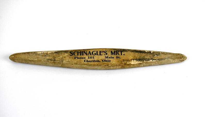 May include: A weathered, elongated wooden object with pointed ends. The text "SCHINAGLE'S MKT." is printed in black, along with address and phone number. The object has a light brown color with signs of wear.