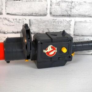 Op de afbeelding: Een zwart en grijs speelgoed protonenpack met een rode nozzle en een handvat. Het speelgoed heeft een Ghostbusters-logo en gele knoppen. Het speelgoed is een replica van het protonenpack uit de film Ghostbusters.
