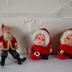 May include: Three vintage Santa Claus figurines. One is standing with a red suit, white beard, and black boots. The other two are sitting with red suits, white fur trim, and white beards.