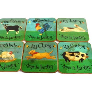 Puede incluir: Juego de seis posavasos cuadrados con ilustraciones pintadas de animales de granja. Cada posavasos presenta un animal diferente: cabra, gallo, conejo, gallina, perro y cerdo. El texto "dans le Jardin" está escrito en cada posavasos.