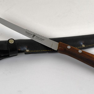 Puede incluir: Un cuchillo de acero inoxidable con borde dentado y mango de madera, sobre una funda de cuero negro. El cuchillo está etiquetado como "Paskelok Silver Eagle".