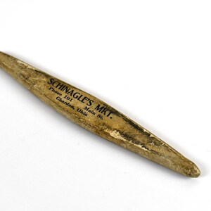 May include: A vintage, elongated wooden object with a pointed end. The object has a light brown color with dark markings and text that reads "SCHINAGLE'S MKT." along with an address and phone number. The item appears to be aged.