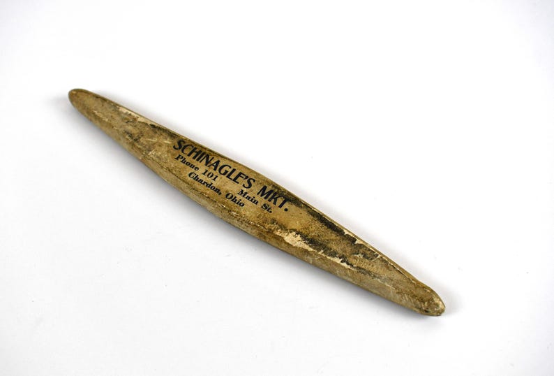 May include: A vintage, elongated wooden object with a pointed end. The object has a light brown color with dark markings and text that reads "SCHINAGLE'S MKT." along with an address and phone number. The item appears to be aged.