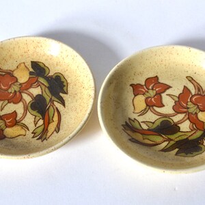 Op de afbeelding: Twee kleine, ronde, gespikkelde gele schaaltjes. Elk schaaltje heeft een bloemenontwerp met rode, oranje, gele en groene bladeren en bloemen. De schaaltjes zijn waarschijnlijk bedoeld voor het serveren van kleine porties of als decoratieve items.