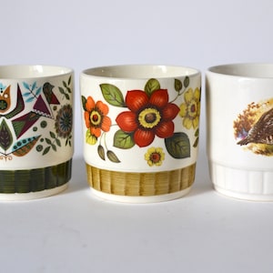 Könnte beinhalten: Drei Keramikbecher mit dekorativen Designs. Ein Becher hat ein Blumenmuster mit roten, orangefarbenen und gelben Blumen. Ein weiterer hat ein Vogel- und Blumenmuster, und der dritte hat eine Vogelillustration.