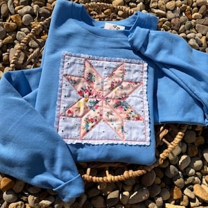 May include: A light blue long-sleeve sweatshirt featuring a square patch on the chest. The patch displays a star pattern made from pink fabric with a floral print, bordered by white eyelet fabric with a scalloped edge and pink stitching.