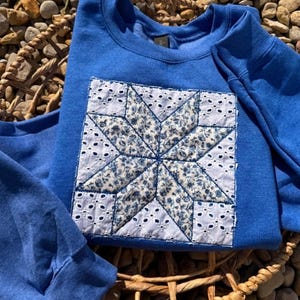 May include: A royal blue long-sleeve sweatshirt featuring a square patch on the chest. The patch displays a star pattern constructed from white eyelet fabric and a blue and white floral print fabric, stitched onto the center of the garment.