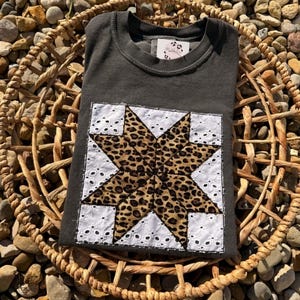 May include: A charcoal gray crew neck t-shirt featuring a square applique patch on the front. The patch displays a large eight-pointed star with a brown and black leopard print pattern, surrounded by white eyelet fabric with a floral cutout design.