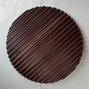May include: A circular, dark brown wooden decorative item featuring a ridged, diagonal pattern. The wooden art piece is mounted on a light grey wall. The ridges create a textured surface, adding visual interest. Likely a wall hanging or decorative plate, it measures approximately 40 cm in diameter.