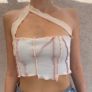 Könnte beinhalten: Ein weißes Crop-Top mit rosa und pfirsichfarbenen Nähten. Das Top hat einen einseitigen Träger und einen rohen Saum.