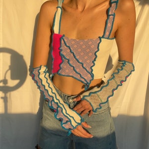 Könnte beinhalten: Ein weißes, rosa und blaues Patchwork-Crop-Top mit Rüschenbesatz. Das Top hat einen quadratischen Ausschnitt und dünne Träger. Das Model trägt das Top zu einer blauen Jeans.