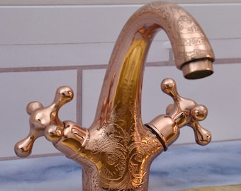 May include: A copper bathroom tap with two cross-shaped handles and an ornate design. The tap is mounted on a white tiled wall above a gold sink.