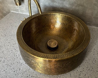 Könnte beinhalten: Ein gehämmertes Messingwaschbecken mit einem verzierten Design am Rand. Das Waschbecken hat einen Abfluss in der Mitte und einen Wasserhahn.