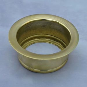 May include: A brass-colored sink drain flange with a circular design. The flange has a wide, flat rim and a cylindrical body. The interior is hollow, and the overall appearance is smooth and metallic.