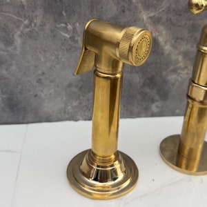 May include: Gold-toned kitchen faucet with a side sprayer. The sprayer has a round nozzle and a lever handle. The faucet has a tall, cylindrical base.