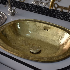 Op de afbeelding: Een ovale gouden badkamer wastafel met een gehamerde afwerking en decoratieve etsen rond de rand. De wastafel staat op een wit aanrecht.