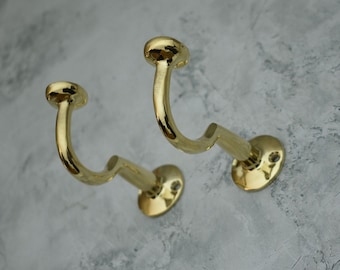 May include: Two gold metal hooks with a rounded top and a curved arm. The hooks are mounted on a wall.