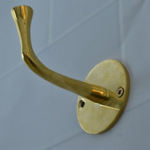 May include: A gold-toned metal hook with a curved arm and a round base. The hook is mounted to a white tiled wall.
