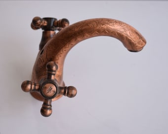May include: A copper faucet with an ornate design and a cross-shaped handle. The faucet has a curved spout and is finished in a warm copper tone.