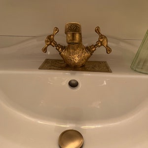 Puede incluir: Un lavabo de baño de porcelana blanca con un grifo y un tapón de desagüe dorados.