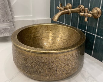 Könnte beinhalten: Ein gehämmertes Messing-Waschbecken mit einem komplizierten floralen Design. Das Waschbecken ist auf einer weißen Arbeitsplatte montiert und hat einen Messinghahn mit Kreuzgriffen.