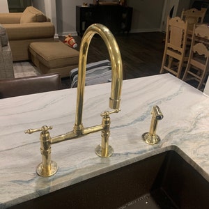 Puede incluir: Un grifo de cocina dorado con un caño de arco alto y dos manijas. El grifo está montado en una encimera blanca con un fregadero marrón.
