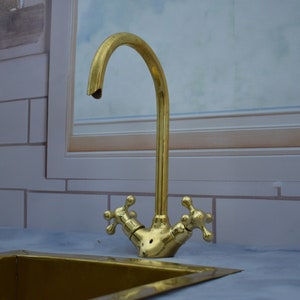 May include: A gold-toned faucet with two cross-shaped handles, mounted on a white tiled countertop. The faucet has a curved spout and is attached to a sink.