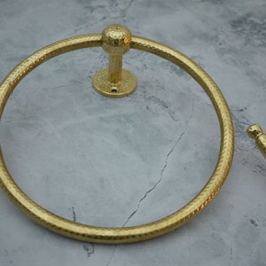 May include: Gold-toned metal towel ring with a hammered finish. The ring is mounted to a wall with a round base.
