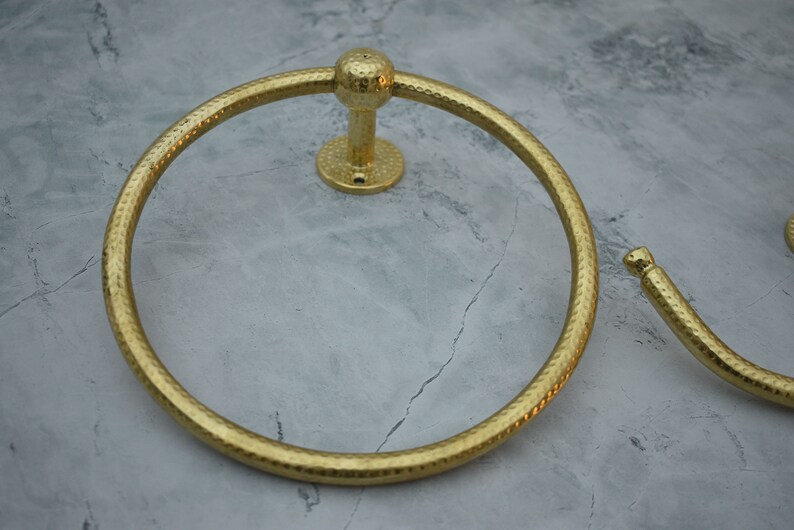 May include: Gold-toned metal towel ring with a hammered finish. The ring is mounted to a wall with a round base.