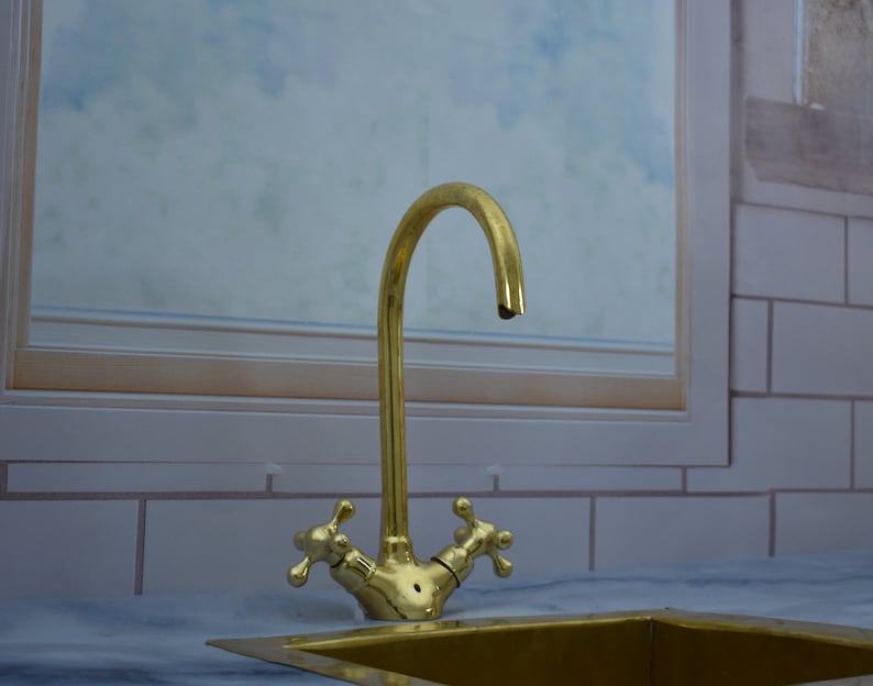 May include: A gold-toned kitchen faucet with two handles and a curved spout. The faucet is mounted on a white countertop.