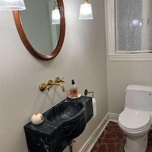 May include: A black marble pedestal sink with a gold faucet and a small white pumpkin on the counter. The sink is in a bathroom with a white toilet and a tiled floor.