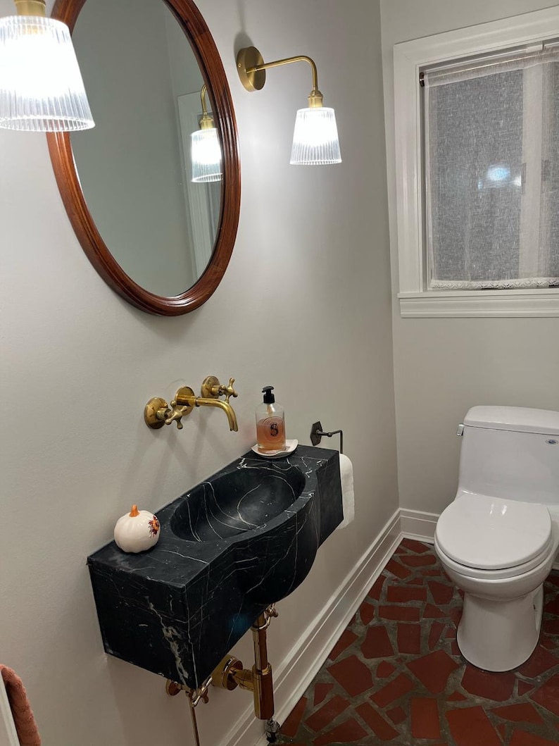 May include: A black marble pedestal sink with a gold faucet and a small white pumpkin on the counter. The sink is in a bathroom with a white toilet and a tiled floor.