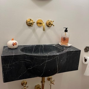 May include: A black marble bathroom sink with a white pumpkin on the counter. The sink has a curved design and gold fixtures. There is a gold soap dispenser with a label that says "S" on the counter.