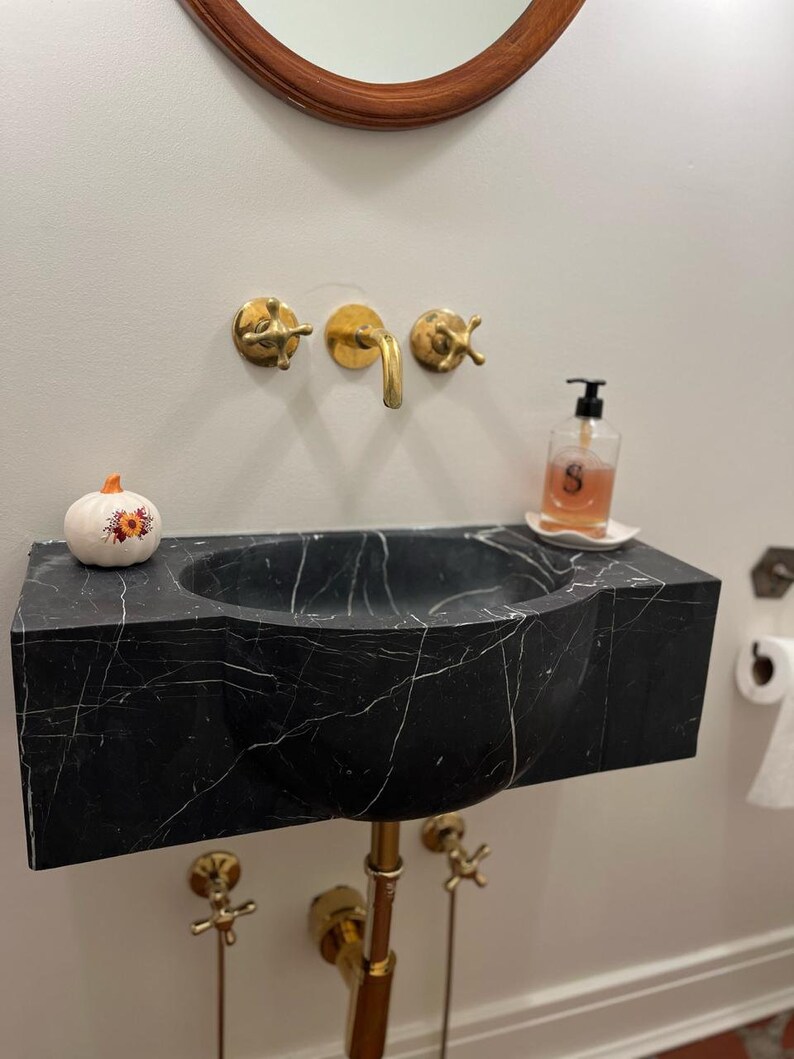 May include: A black marble bathroom sink with a white pumpkin on the counter. The sink has a curved design and gold fixtures. There is a gold soap dispenser with a label that says "S" on the counter.