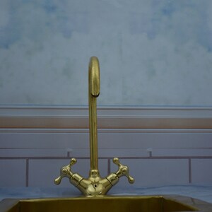 May include: A gold-toned faucet with two handles, mounted on a white tiled wall. The faucet is in front of a window with a blurry background.