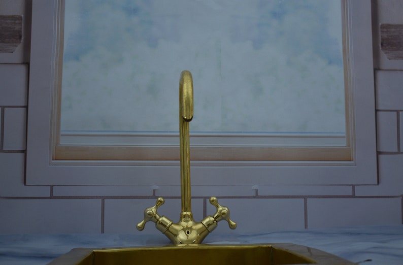 May include: A gold-toned faucet with two handles, mounted on a white tiled wall. The faucet is in front of a window with a blurry background.