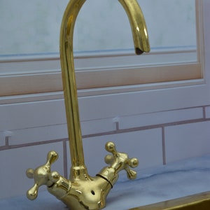 May include: A gold-toned faucet with two cross-shaped handles. The faucet has a curved spout and is mounted on a countertop.
