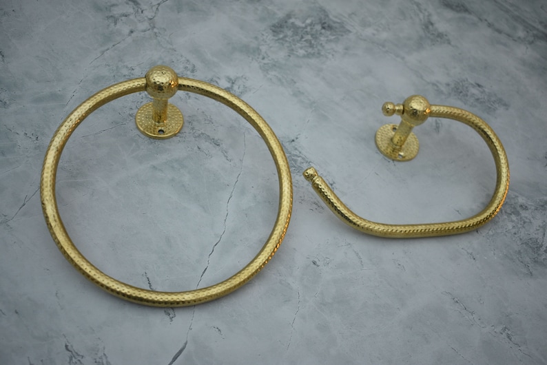 May include: Two gold-toned towel rings with a hammered finish. The larger ring is circular, while the smaller ring is a curved shape.