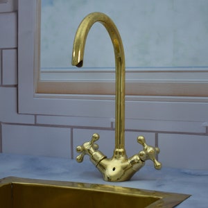 May include: A gold-toned kitchen faucet with a high-arc spout and two cross-shaped handles. The faucet is mounted on a white countertop.