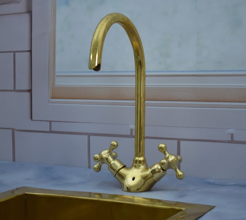 May include: A gold-toned kitchen faucet with a high-arc spout and two cross-shaped handles. The faucet is mounted on a white countertop.