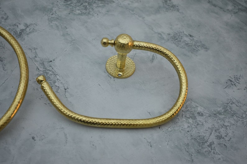 May include: Two gold-toned metal towel rings with a hammered texture. The rings are attached to a wall with a round base.