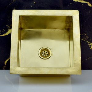 May include: A square brass sink with a drain in the centre. The sink is set against a black background.